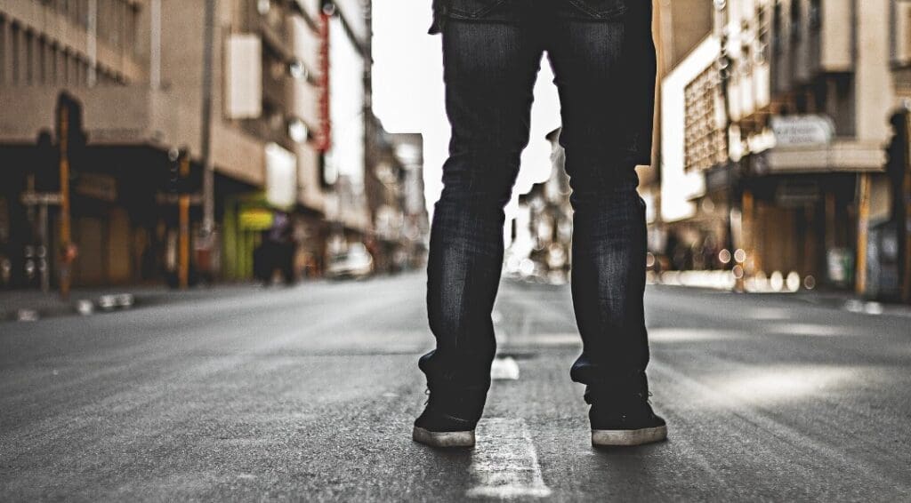 A person standing in the middle of a deserted city street. The view is from behind, focusing on their legs and feet, with a blurred background of buildings on both sides of the street.