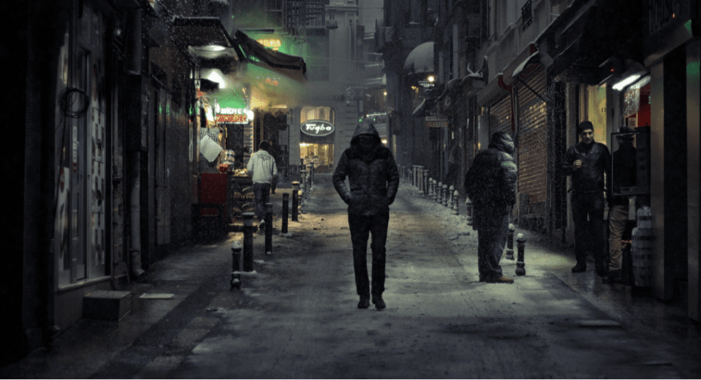 A person in a dark hooded jacket walking down a narrow, snow-covered street in the evening, surrounded by buildings and a few other people.