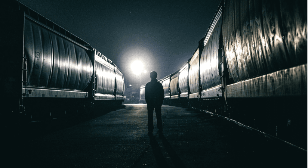 A silhouette of a person standing alone between two long rows of train cars at night, with a bright light shining directly ahead.