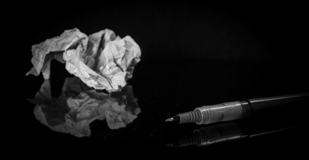 A crumpled piece of paper and a pen lying on a reflective black surface, set against a dark background.