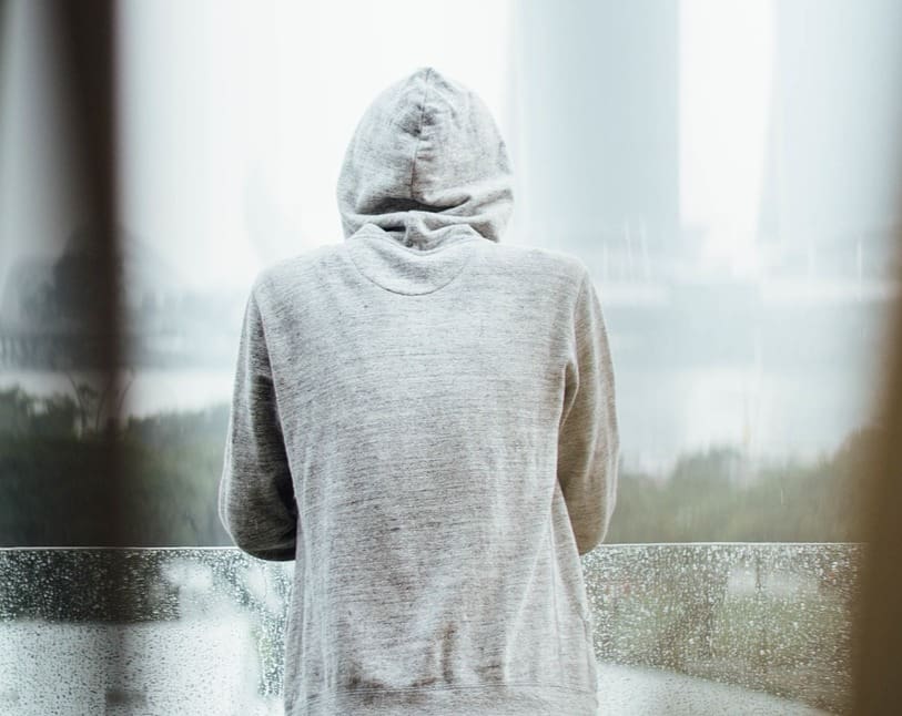 Person in a gray hoodie standing with their back turned, looking out over a misty, blurred cityscape.