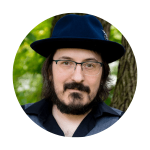 Profile photo of Frank Tybush V, an author. He is wearing a dark blue hat and glasses, with a short beard and mustache. He is standing outdoors with a tree and greenery visible in the background.