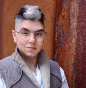 A headshot of Carlin Thomas, a person with short, black-and-white dyed hair, wearing round glasses and a textured gray vest over a white shirt. They are standing against a rust-colored, textured metal background, giving the photo an industrial and modern feel.