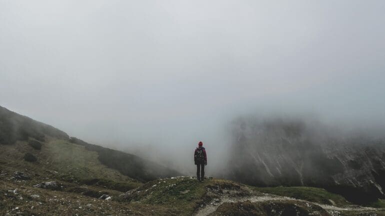 person standing on a grassy patch of a hill or mountain overcome with fog and clouds