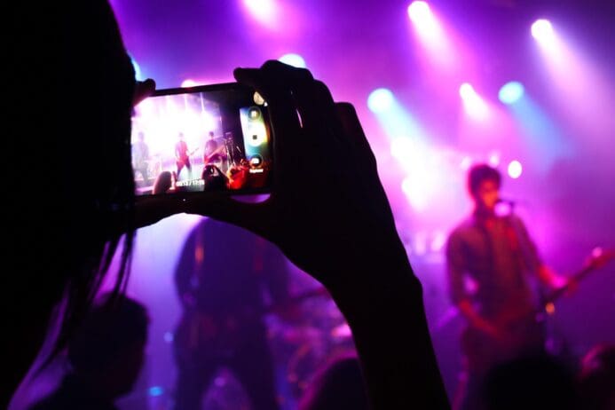 someone recording a concert on their smartphone there is a 4 person band and lots of colorful lights and lots of purples