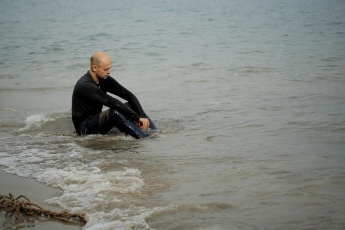 man laying in the sand on the shoreline looking pensively at the water while sitting in it