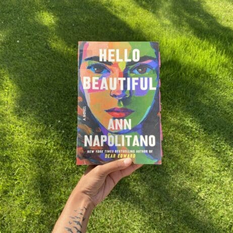 A hand holding up the book "Hello Beautiful" by Ann Napolitano against a backdrop of green grass. The book cover features a vibrant, multicolored illustration of a woman's face, with the title and author's name in bold white text. The hand holding the book has a small tattoo, and the overall scene is set outdoors, with the sunlight casting soft shadows on the grass.