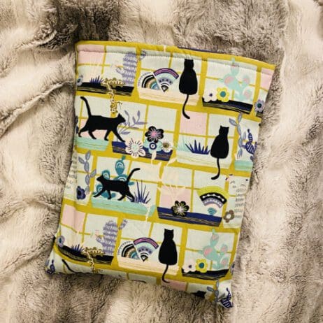 The image shows a padded fabric cover or pouch, likely designed for a tablet or book, lying on a soft, gray fur-like surface. The fabric features a whimsical pattern with black silhouettes of cats in various poses, along with colorful flowers, plants, and abstract designs. The background of the fabric is a mix of pastel colors, including pink, yellow, and blue, creating a playful and cozy aesthetic.