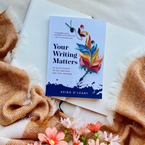 The image shows the book "Your Writing Matters" by Keiko O'Leary placed on top of an open notebook with dotted pages. The book cover features a vibrant quilled paper design of a feather in various colors, with the subtitle "34 Quick Essays to Get Unstuck and Stay Inspired" beneath the main title. Surrounding the book is a soft, fringed, brown and white blanket, and pink flowers are partially visible at the bottom of the image, creating a cozy and inviting scene.