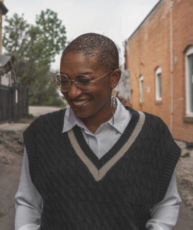 Author photo of Dami Salako smiling, wearing glasses, a collared shirt, and a dark knitted vest, with a background of an outdoor scene with trees and a brick building.