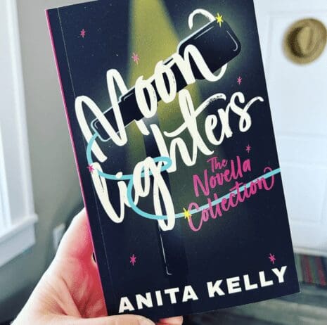 The image shows a person holding a book titled "Moonlighters: The Novella Collection" by Anita Kelly. The cover features a dark background with the title written in large, playful white and pink script fonts. A spotlight illustration shines on the word "Moonlighters," and a blue ribbon-like design wraps around the title. The author's name is displayed at the bottom in bold white letters. The background suggests an indoor setting, with part of a door and a wall-mounted hat visible.
