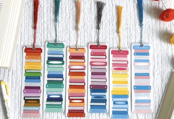 The image shows a set of six colorful bookmarks laid out on a white textured surface. Each bookmark features a unique pattern of stylized book spines stacked vertically in various colors, including red, blue, yellow, pink, and green. Each bookmark has a tassel at the top, with the tassels in different colors matching the design of the bookmarks. Surrounding the bookmarks are parts of an open book, a yellow and white pen, and some decorative elements like colorful pom-poms. The scene is bright and visually appealing, perfect for book enthusiasts.