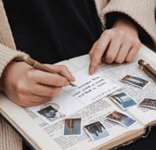 A close-up of a person writing in a journal with a fountain pen. The journal is open on a page filled with text, and several small, printed photos or stickers are scattered across the pages. The person is wearing a cozy beige sweater, suggesting a relaxed and thoughtful moment of reflection or creativity.