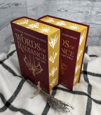 The image shows two hardcover volumes of "Words of Radiance" by Brandon Sanderson, part of "The Stormlight Archive" series. The books are placed on a black and white checkered blanket. Both volumes have a rich burgundy cover with gold accents, and the title is prominently displayed in gold lettering. The edges of the pages are gilded with intricate white designs, adding a luxurious touch. A small bundle of dried lavender is placed in front of the books, contributing to the cozy and elegant atmosphere of the scene.
