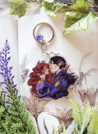 A detailed illustration of a keychain featuring two fantasy characters in a romantic pose. The characters, one with long red hair and animal-like ears and the other with dark hair and pointed elf-like ears, are depicted in rich, vibrant colors. The keychain is placed on an open book with botanical sketches, surrounded by artificial greenery and purple flowers, adding a natural and whimsical atmosphere to the scene.