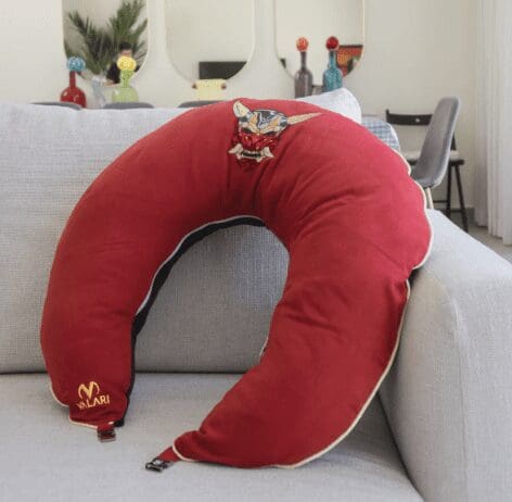 A red crescent-shaped pillow with an embroidered design of a fierce, stylized face, possibly resembling a dragon or demon, at the top center. The pillow is placed on a light gray sofa in a modern living room. The brand name 'VILARI' is embroidered in gold near the bottom left corner of the pillow. The room features minimalist decor, with colorful glass bottles on a mirrored shelf in the background and a chair visible to the right.