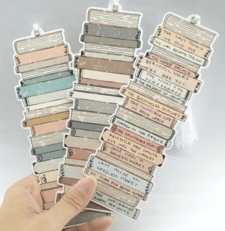 The image shows a hand holding three decorative bookmarks shaped like stacked books. Each bookmark features a stack of illustrated books in muted colors like pastel pinks, greens, and grays. The spines of the books have various titles written on them, resembling handwritten text. The bookmarks have a tassel at the top, and the overall design is whimsical and charming, perfect for book lovers.
