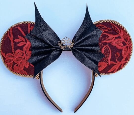 A decorative headband featuring large, round ears covered in red fabric with an intricate floral pattern, trimmed with gold rope. The center of the headband is adorned with a black, bat-shaped bow made of a glittery material, with a small gold lotus flower embellishment at the center. The design combines elegant and gothic elements, creating a unique and stylish accessory.