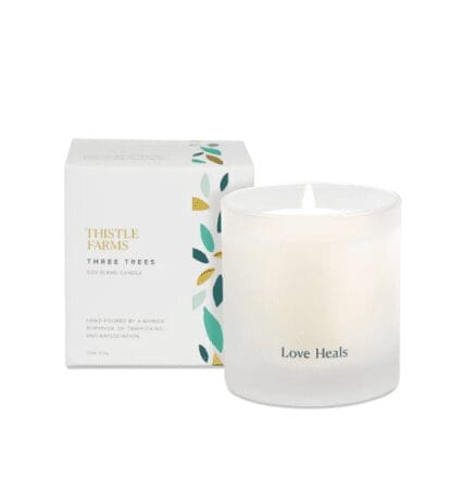 A white soy blend candle from Thistle Farms with the phrase "Love Heals" printed on the front of the frosted glass container. The candle is lit, creating a warm glow. Behind the candle is the product box, which is white with a minimalist design featuring leaves and the text "Thistle Farms Three Trees Soy Blend Candle." The box also notes that the candle is hand-poured by a woman surviving trafficking and exploitation.