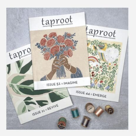The image shows three issues of "Taproot" magazine arranged in a fan-like layout on a gray surface. The magazine covers feature artistic illustrations. The central issue, "Issue 51 :: IMAGINE," has an illustration of hands holding a bouquet of red flowers. To the left, "Issue 31 :: REVIVE" displays abstract green foliage, and to the right, "Issue 44 :: EMERGE" features a colorful scene with animals, plants, and a rainbow. Below the magazines are several small spools of thread in various colors.