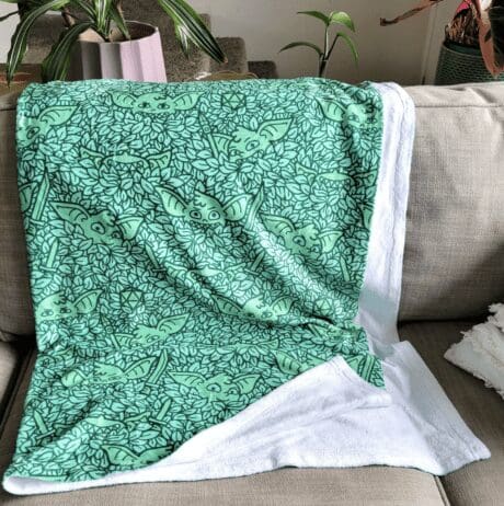 A soft, green blanket with a playful pattern featuring outlines of Baby Yoda's face peeking through leaves. The blanket is draped over a beige sofa, with the white, fluffy underside slightly exposed. In the background, there are a few potted plants and a staircase, creating a cozy, inviting living space.