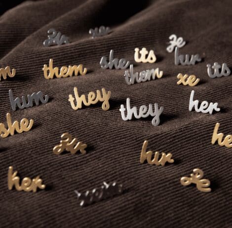A collection of small metallic pins, each displaying different pronouns in cursive script, spread across a textured, dark brown fabric surface. The pronouns include 'they,' 'them,' 'she,' 'her,' 'he,' 'him,' 'xe,' 'ze,' 'its,' 'hir,' 'hum,' and others, in both gold and silver finishes. The pins are scattered randomly, creating a visually appealing arrangement that emphasizes inclusivity and diverse gender identities.