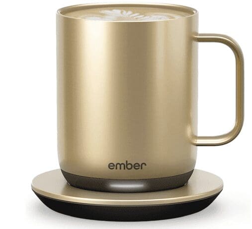A gold-colored Ember temperature-controlled mug sitting on its matching charging coaster. The mug has a sleek, modern design with the brand name "ember" printed near the base. The top of the mug shows a latte with art in the foam.