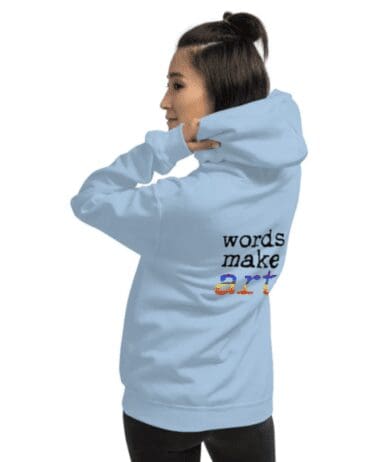 The image shows a person wearing a light blue hoodie with the text "words make art" printed on the back. The words "words make" are in black, and the word "art" is in a gradient of rainbow colors. The person is facing away from the camera and has their right hand near their face, slightly pulling the hoodie.