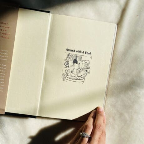 Photo of a person reading a book that the inside cover has the words Armed with A Book and a lovely illustration of someone reading on a big cheer sitting criss-cross with a lovely bookshelf behind them and a cat, it is a line drawing in black.
