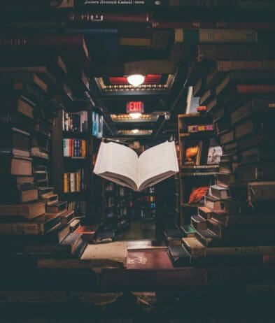 A visually captivating scene inside a bookstore, with an open book seemingly floating in the air at the center of the image. The book is framed by an archway made of stacked books, creating a tunnel-like effect that draws the viewer's eye towards the book. The background reveals rows of shelves filled with more books, under warm lighting, contributing to the cozy and magical atmosphere. A red "EXIT" sign is visible in the distance, adding a touch of contrast to the scene.